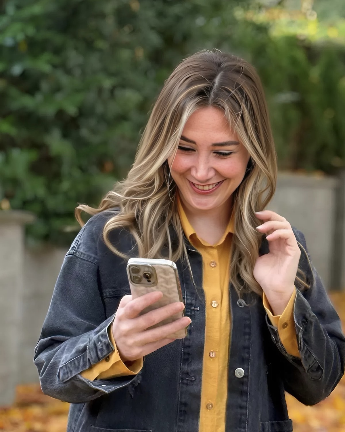 Sara Gholamali Poor smiling at her phone, in a dark denim jacket and mustard shirt, with autumn leaves behind her.